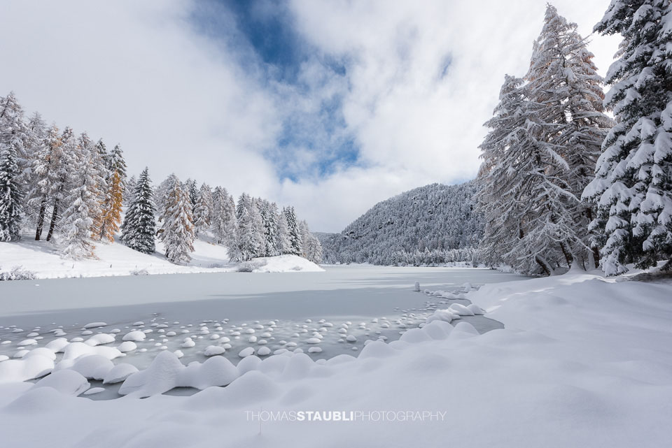 Der zugefrorene Lai da Palpuogna unter einer aufreissenden Wolkendecke, umgeben von schneebedeckten Lärchen und leuchtend orangefarbenem Herbstlaub.