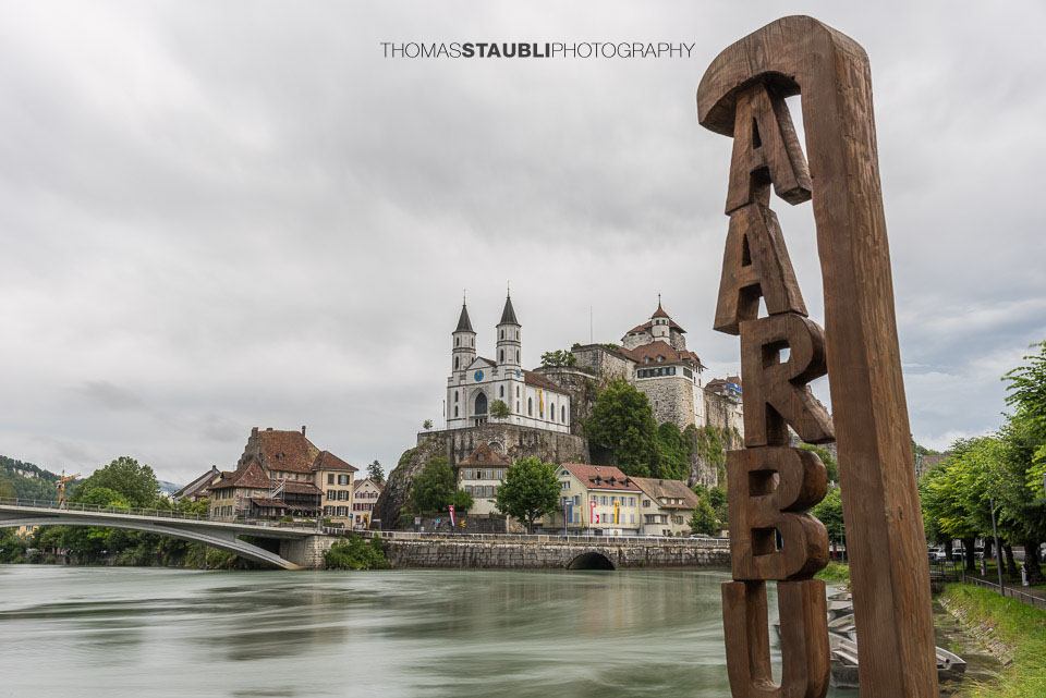 Im Vordergrund steht ein grosser hölzerner Schriftzug «AARBURG», dahinter die markante Festung und reformierte Kirche auf einem Felsen über der Aare.