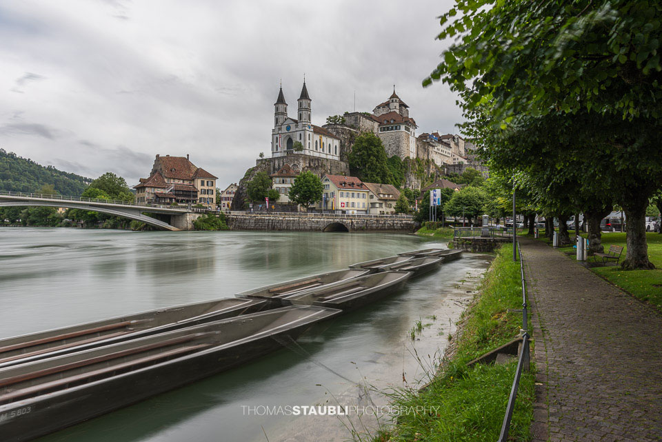 Die Festung Aarburg und die reformierte Kirche erheben sich eindrucksvoll über dem Fluss, während im Vordergrund mehrere Weidlinge am ruhigen Ufer liegen.