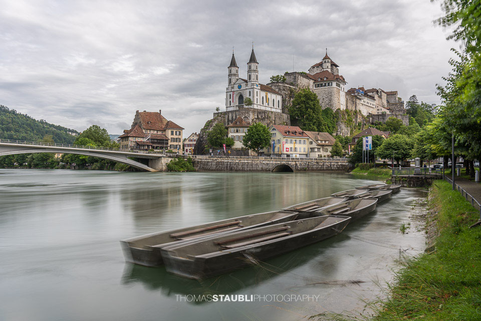 Die Festung Aarburg und die reformierte Kirche erheben sich eindrucksvoll über dem Fluss, während im Vordergrund mehrere Weidlinge am ruhigen Ufer liegen.