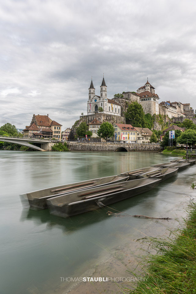 Die Festung Aarburg und die reformierte Kirche erheben sich eindrucksvoll über dem Fluss, während im Vordergrund mehrere Weidlinge am ruhigen Ufer liegen.