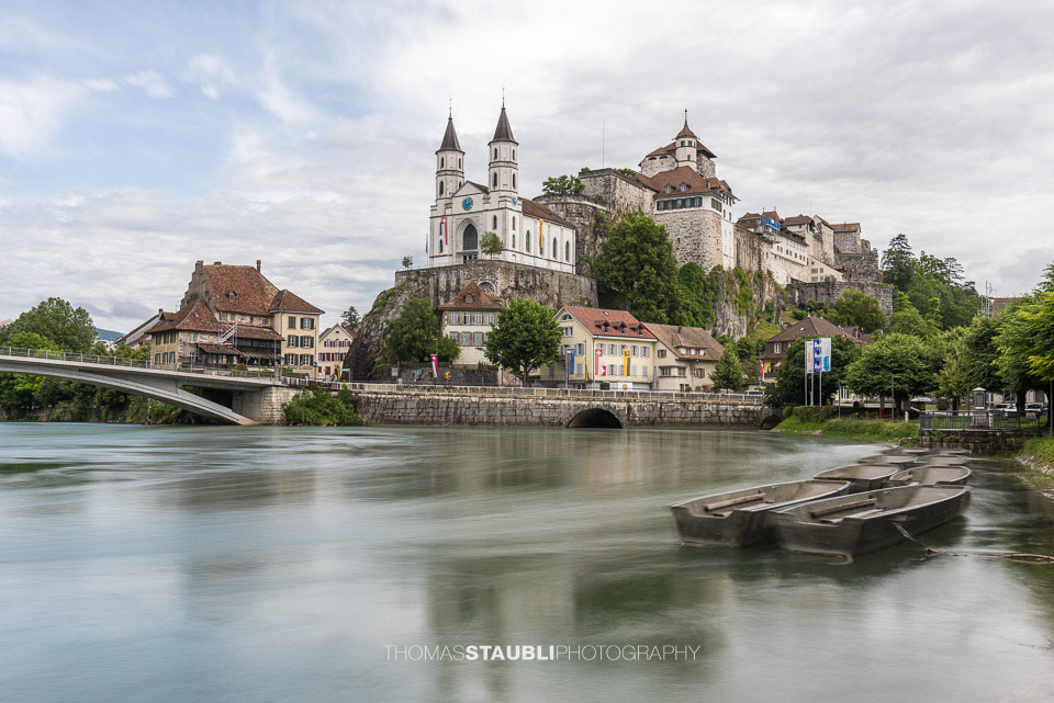 Die Festung Aarburg und die reformierte Kirche erheben sich eindrucksvoll über dem Fluss, während im Vordergrund mehrere Weidlinge am ruhigen Ufer liegen.