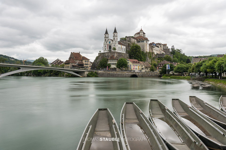 Die Festung Aarburg und die reformierte Kirche erheben sich eindrucksvoll über dem Fluss, während im Vordergrund mehrere Pontonier Boote am ruhigen Ufer liegen.
