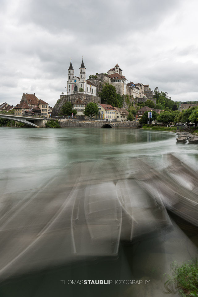 Die Festung Aarburg und die reformierte Kirche erheben sich eindrucksvoll über dem Fluss, während im Vordergrund mehrere Weidlinge am ruhigen Ufer liegen.