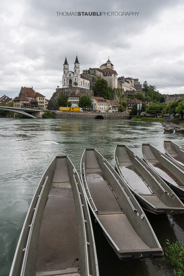 Die Festung Aarburg und die reformierte Kirche erheben sich eindrucksvoll über dem Fluss, während im Vordergrund mehrere Pontonier Boote am ruhigen Ufer liegen.