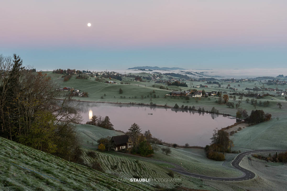 Hügellandschaft mit Raureif bei Vollmond, ein stiller Hüttnersee spiegelt das Mondlicht wider, umgeben von Wiesen und Häusern