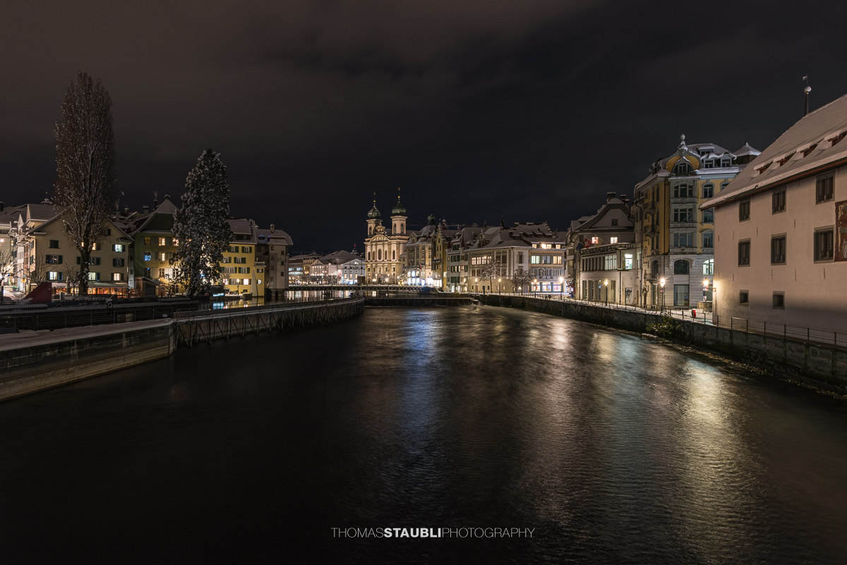 Nächtlicher Blick auf die Jesuitenkirche in Luzern, umgeben von beleuchteten historischen Gebäuden und der Reuss im Vordergrund