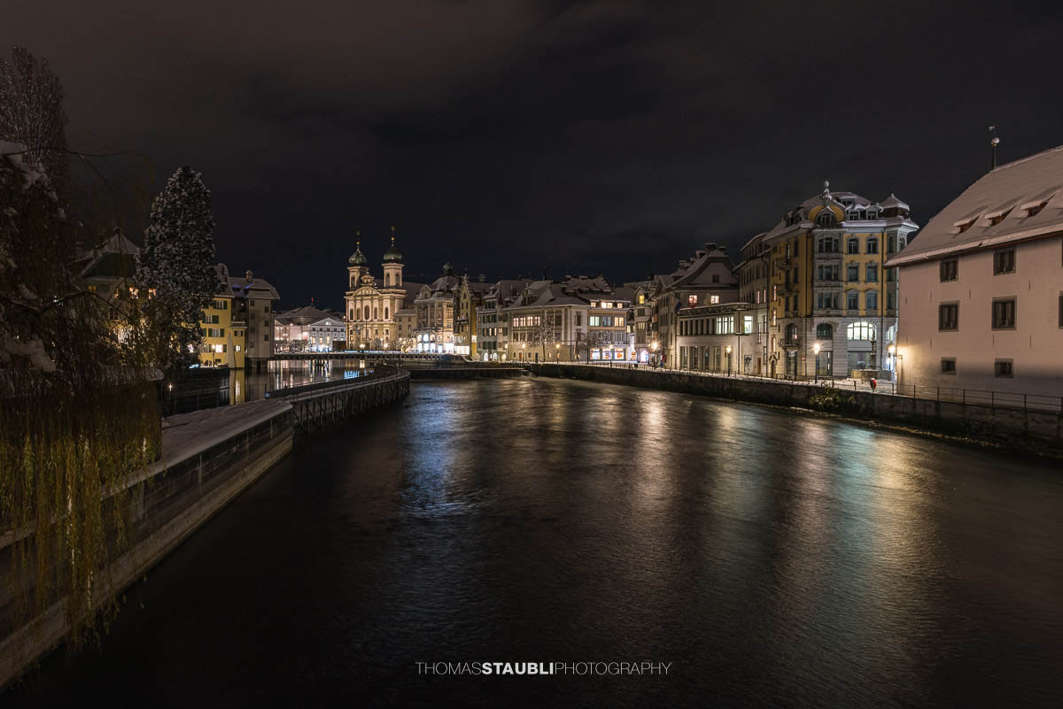 Nächtlicher Blick auf die Jesuitenkirche in Luzern, umgeben von beleuchteten historischen Gebäuden und der Reuss im Vordergrund