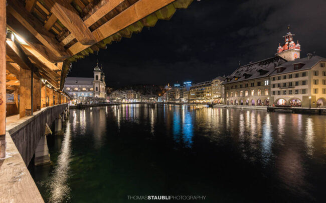 Abendlicher Blick von der Kapellbrücke in Luzern mit erleuchteten Fassaden, der Jesuitenkirche und der Reuss im Vordergrund