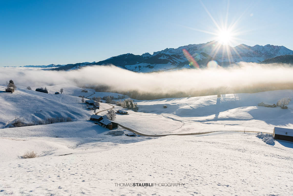 Winterlandschaft mit Schnee bedeckten Hügeln, Nebel im Tal und Bergen im Sonnenaufgang, durchzogen von einer geschwungenen Strasse und verstreuten Höfen.