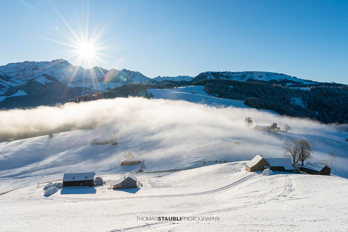 Winterlandschaft mit Schnee bedeckten Hügeln, Nebel im Tal und Bergen im Sonnenaufgang, durchzogen von einer geschwungenen Strasse und verstreuten Höfen.