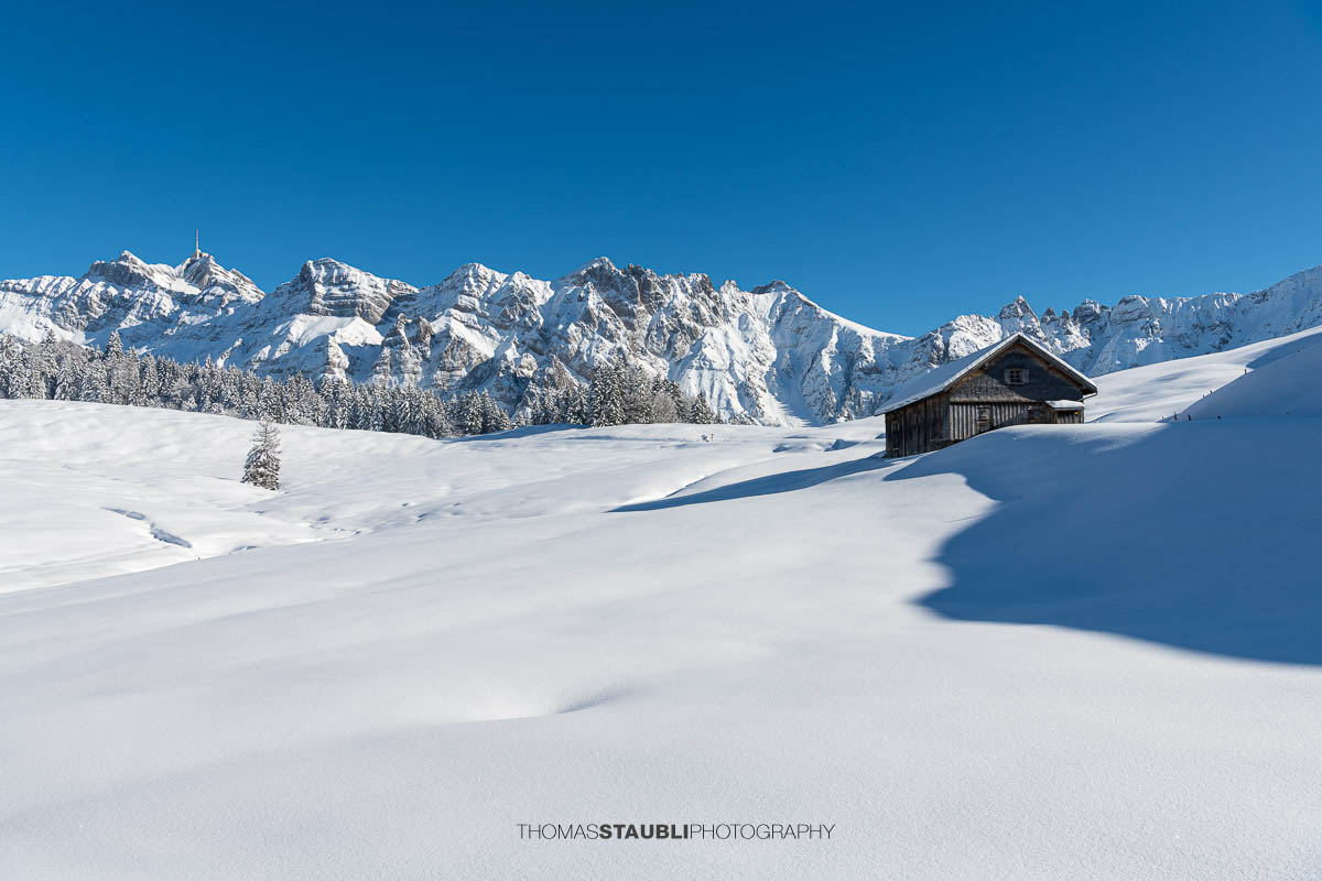 Winterliche Berghütte mit Säntis im Hintergrund unter strahlend blauem Himmel