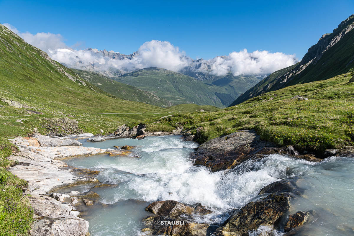 Die Witenwasserenreuss rauscht über Felsstufen durch das grüne Witenwasserental im Kanton Uri, im Hintergrund das Panorama der Urner Alpen unter einem klaren Himmel.