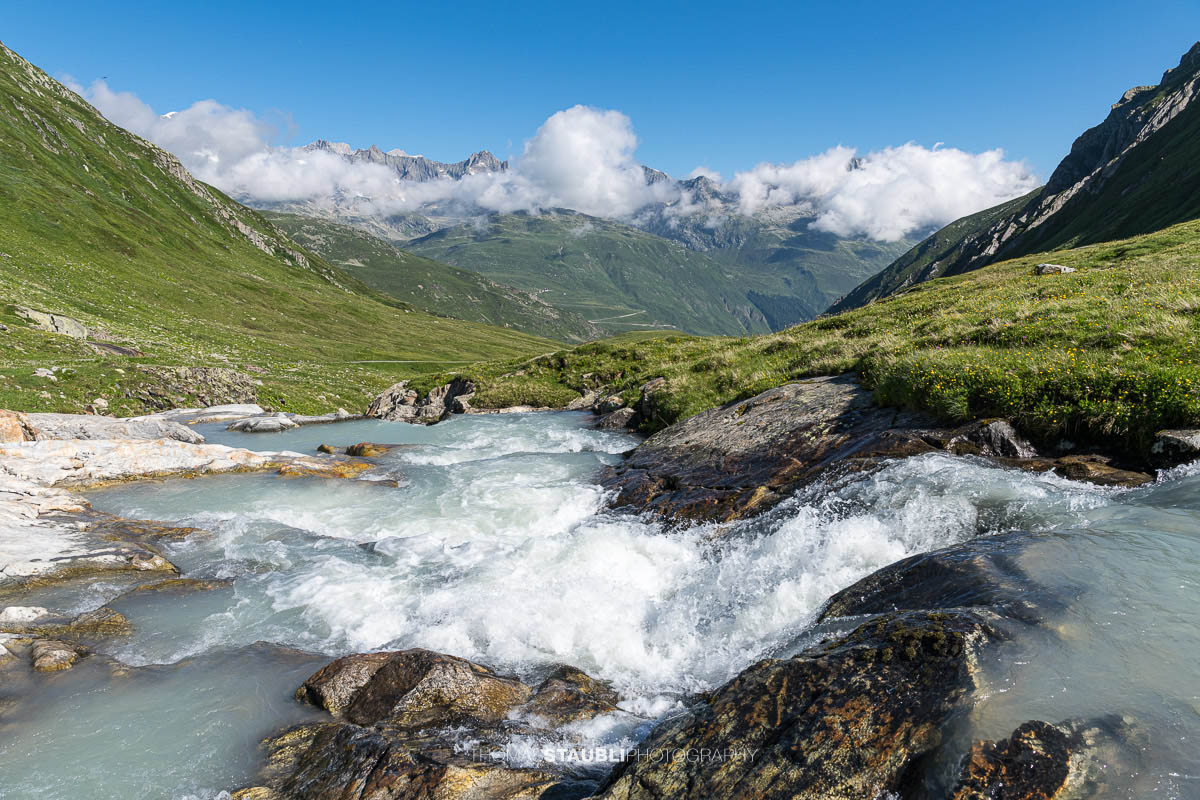 Die Witenwasserenreuss rauscht über Felsstufen durch das grüne Witenwasserental im Kanton Uri, im Hintergrund das Panorama der Urner Alpen unter einem klaren Himmel.
