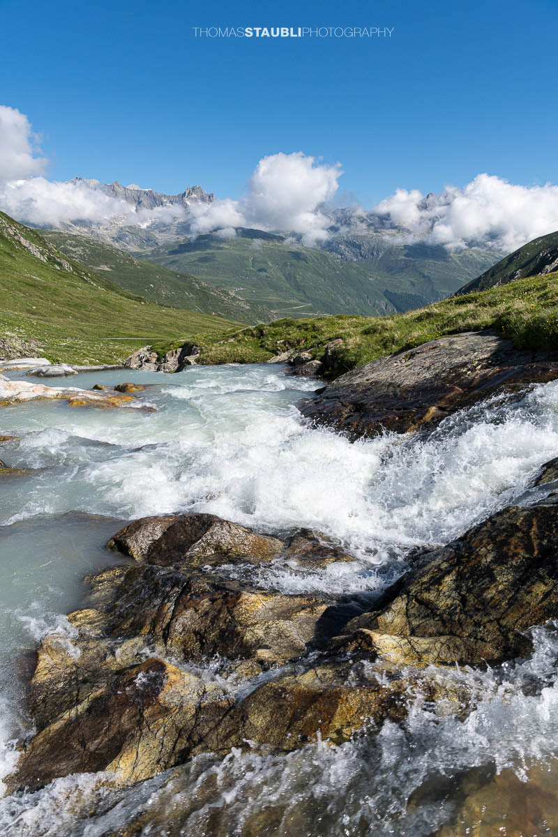 Die Witenwasserenreuss rauscht über Felsstufen durch das grüne Witenwasserental im Kanton Uri, im Hintergrund das Panorama der Urner Alpen unter einem klaren Himmel.