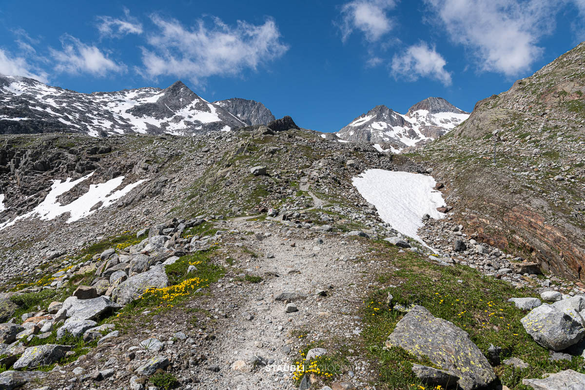 Ein alpiner Wanderweg führt über Geröll und Schneefelder durch die hochalpine Landschaft des Witenwasserentals, mit Blick auf die Gipfel rund um den Witenwasserenstock.