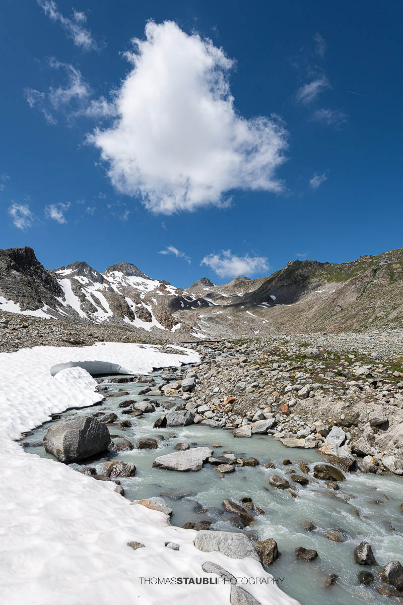 Blick auf die schmelzende Witenwasserenreuss, die sich durch das Geröllfeld schlängelt. Im Hintergrund erheben sich die Schneefelder und Gipfel rund um das Witenwasserental unter einem strahlend blauen Himmel mit vereinzelten Wolken.