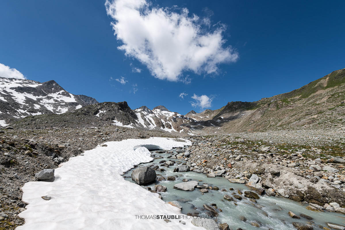 Blick auf die schmelzende Witenwasserenreuss, die sich durch das Geröllfeld schlängelt. Im Hintergrund erheben sich die Schneefelder und Gipfel rund um das Witenwasserental unter einem strahlend blauen Himmel mit vereinzelten Wolken.
