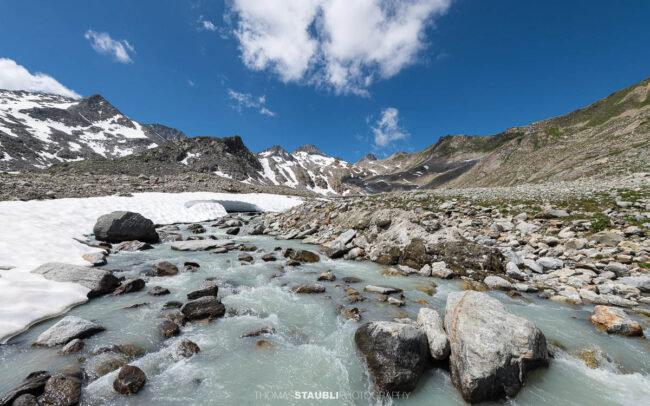 Blick auf die schmelzende Witenwasserenreuss, die sich durch das Geröllfeld schlängelt. Im Hintergrund erheben sich die Schneefelder und Gipfel rund um das Witenwasserental unter einem strahlend blauen Himmel mit vereinzelten Wolken.