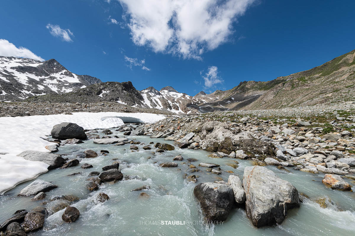 Blick auf die schmelzende Witenwasserenreuss, die sich durch das Geröllfeld schlängelt. Im Hintergrund erheben sich die Schneefelder und Gipfel rund um das Witenwasserental unter einem strahlend blauen Himmel mit vereinzelten Wolken.