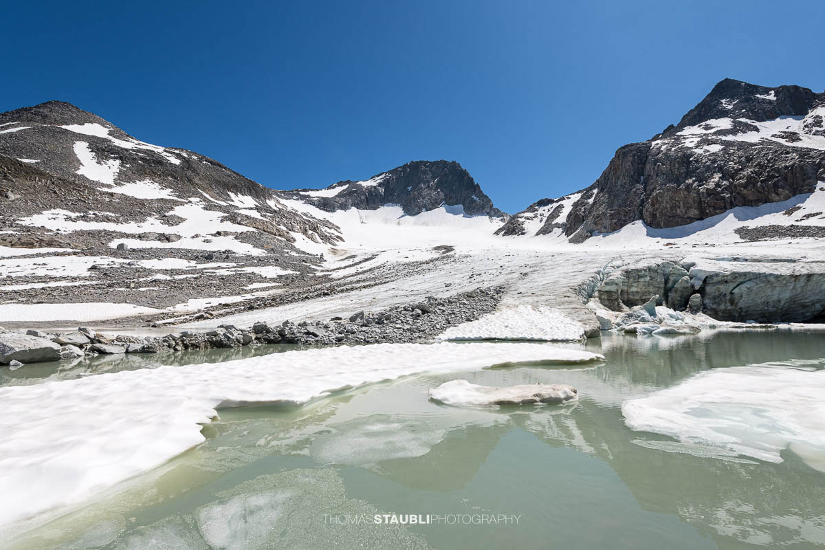 Der Witenwasserengletscher mit seiner Gletscherzunge, die in einen kleinen türkisfarbenen Gletschersee übergeht. Eisschollen treiben im Schmelzwasser, während steile Felswände die Szene rahmen.