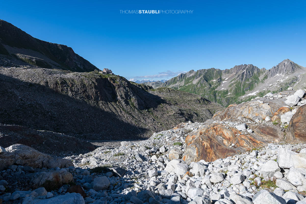 Die Rotondohütte thront auf einem Felsvorsprung oberhalb des Witenwasserentals. Im Vordergrund liegt ein karges Geröllfeld, dahinter erstrecken sich grüne Bergflanken und felsige Gipfel unter einem klaren Himmel.