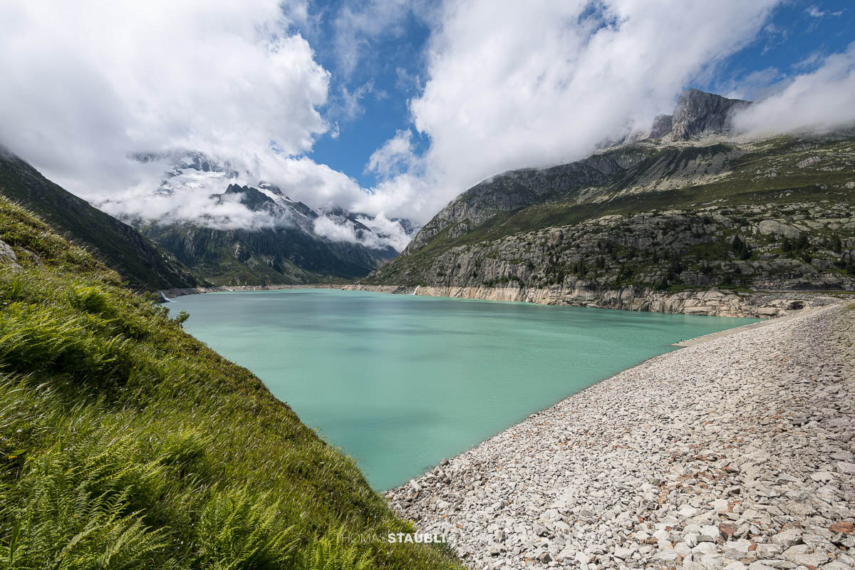 Türkisblauer Göscheneralpsee im Göschenertal, mit Blick ins Chelenalptal und umgeben von steilen Felswänden und Wolken über den Berggipfeln im Kanton Uri.
