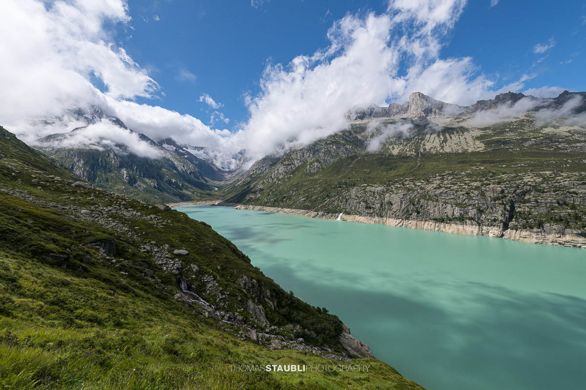 Türkisblauer Göscheneralpsee im Göschenertal, mit Blick ins Chelenalptal und umgeben von steilen Felswänden und Wolken über den Berggipfeln im Kanton Uri.