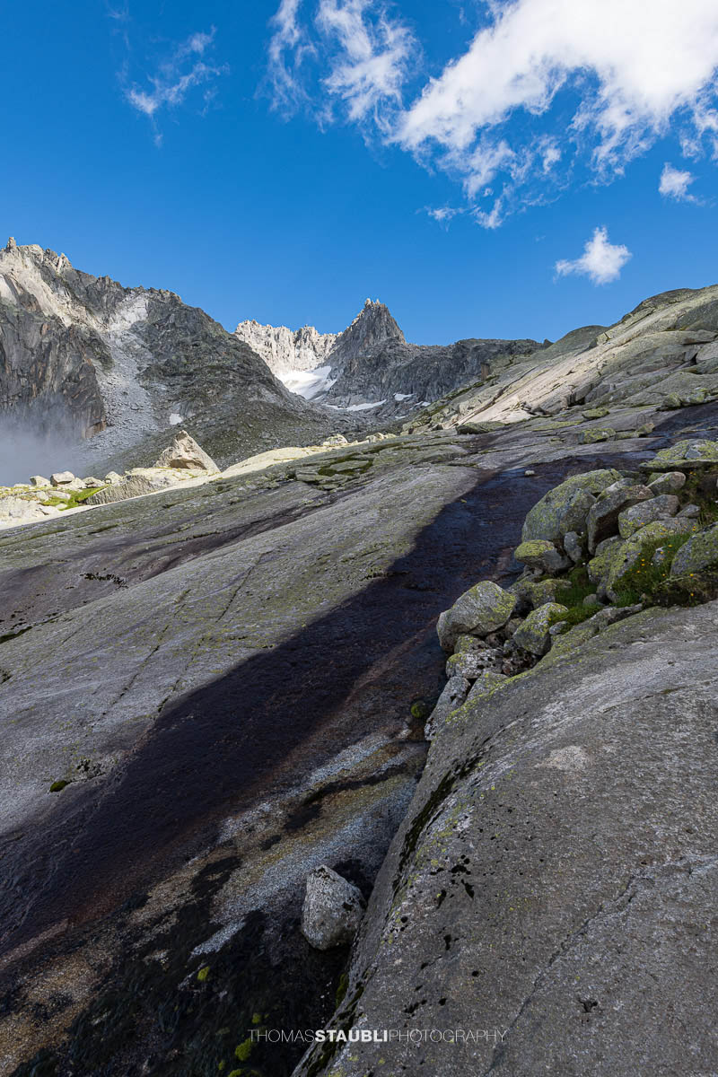 Felsige Älprigenplatten im Vordergrund, dahinter der spitze Gipfel des Feldschijen, teilweise von Nebelschwaden umgeben, unter blauem Himmel in den Urner Alpen.