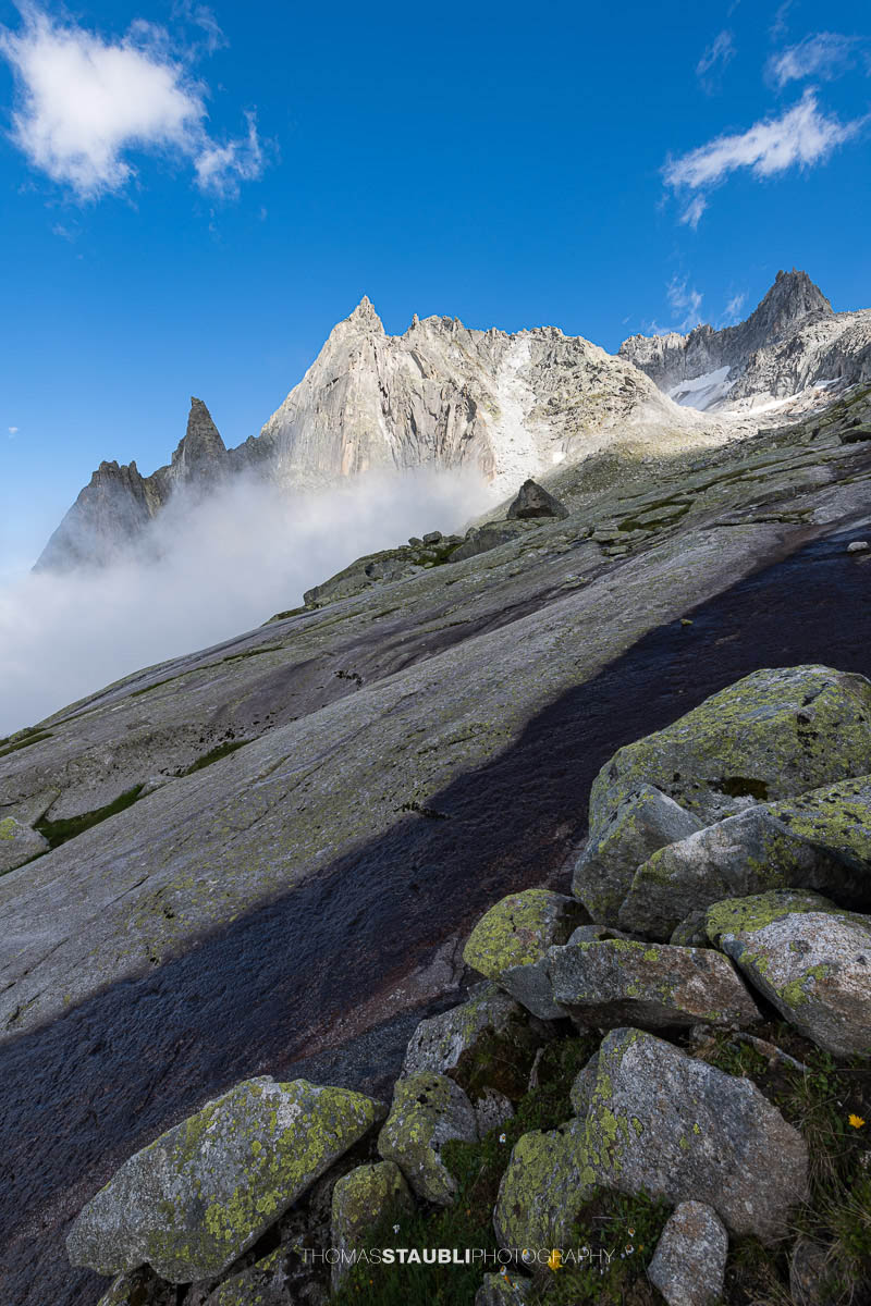 Felsige Älprigenplatten im Vordergrund, dahinter der spitze Gipfel des Feldschijen, teilweise von Nebelschwaden umgeben, unter blauem Himmel in den Urner Alpen.