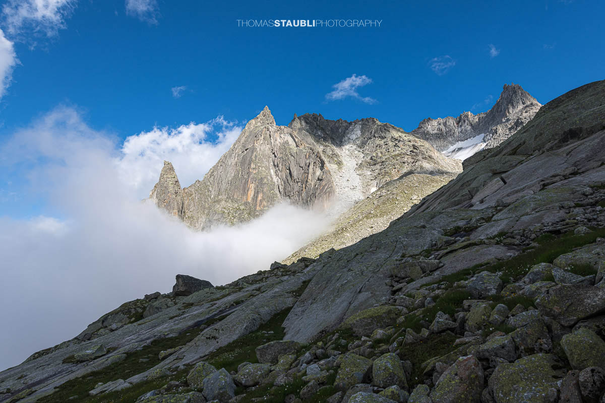 Felsige Älprigenplatten im Vordergrund, dahinter der spitze Gipfel des Feldschijen, teilweise von Nebelschwaden umgeben, unter blauem Himmel in den Urner Alpen.
