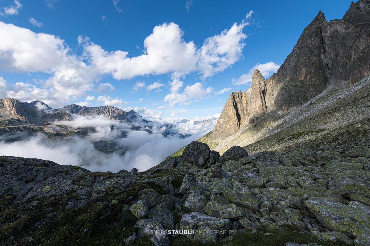 Felsige Landschaft der Älprigenplatten mit Aussicht ins Göschenertal, von Nebelschwaden durchzogen, umgeben von schroffen Gipfeln unter blauem Himmel.