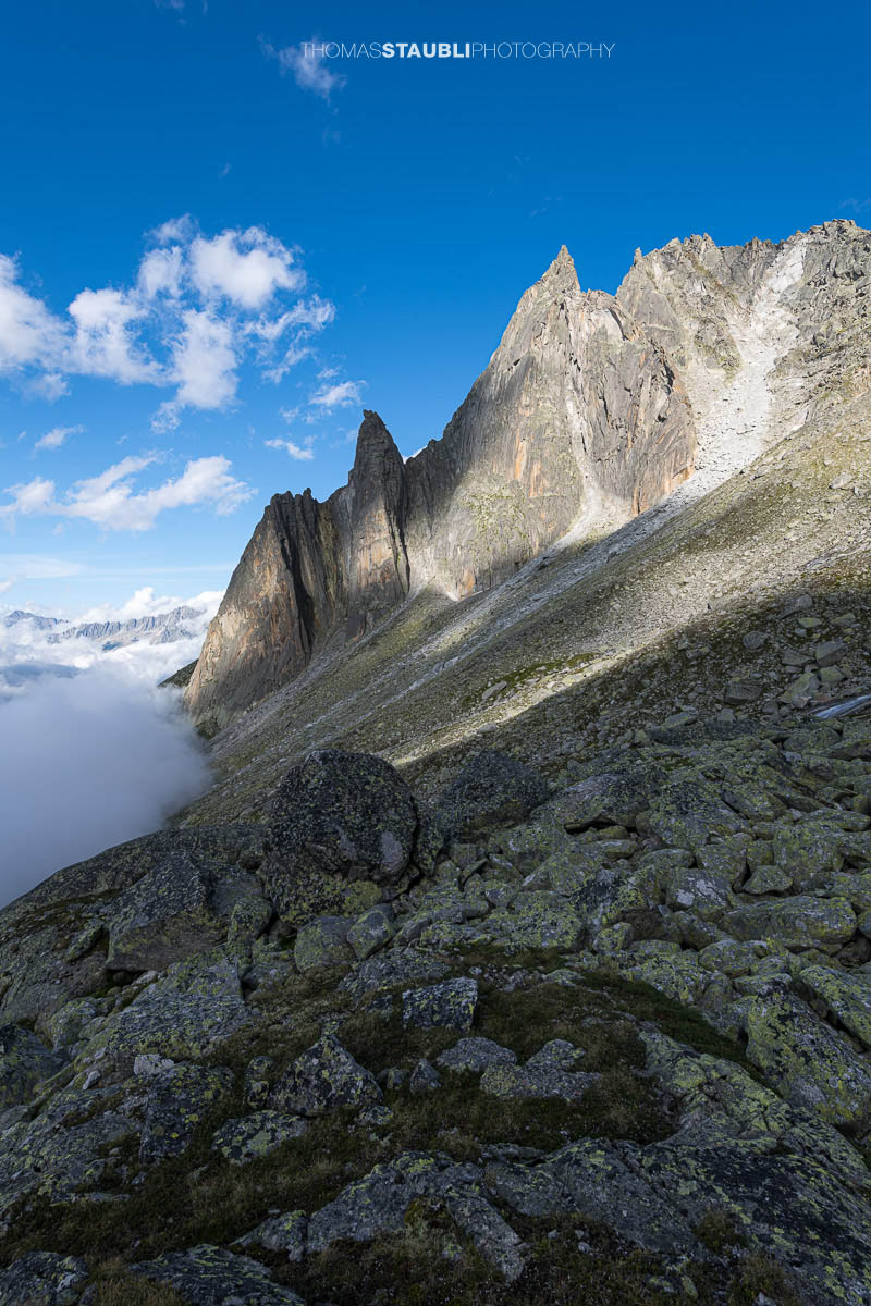 Felsige Älprigenplatten im Vordergrund, dahinter der spitze Gipfel des Feldschijen, teilweise von Nebelschwaden umgeben, unter blauem Himmel in den Urner Alpen.