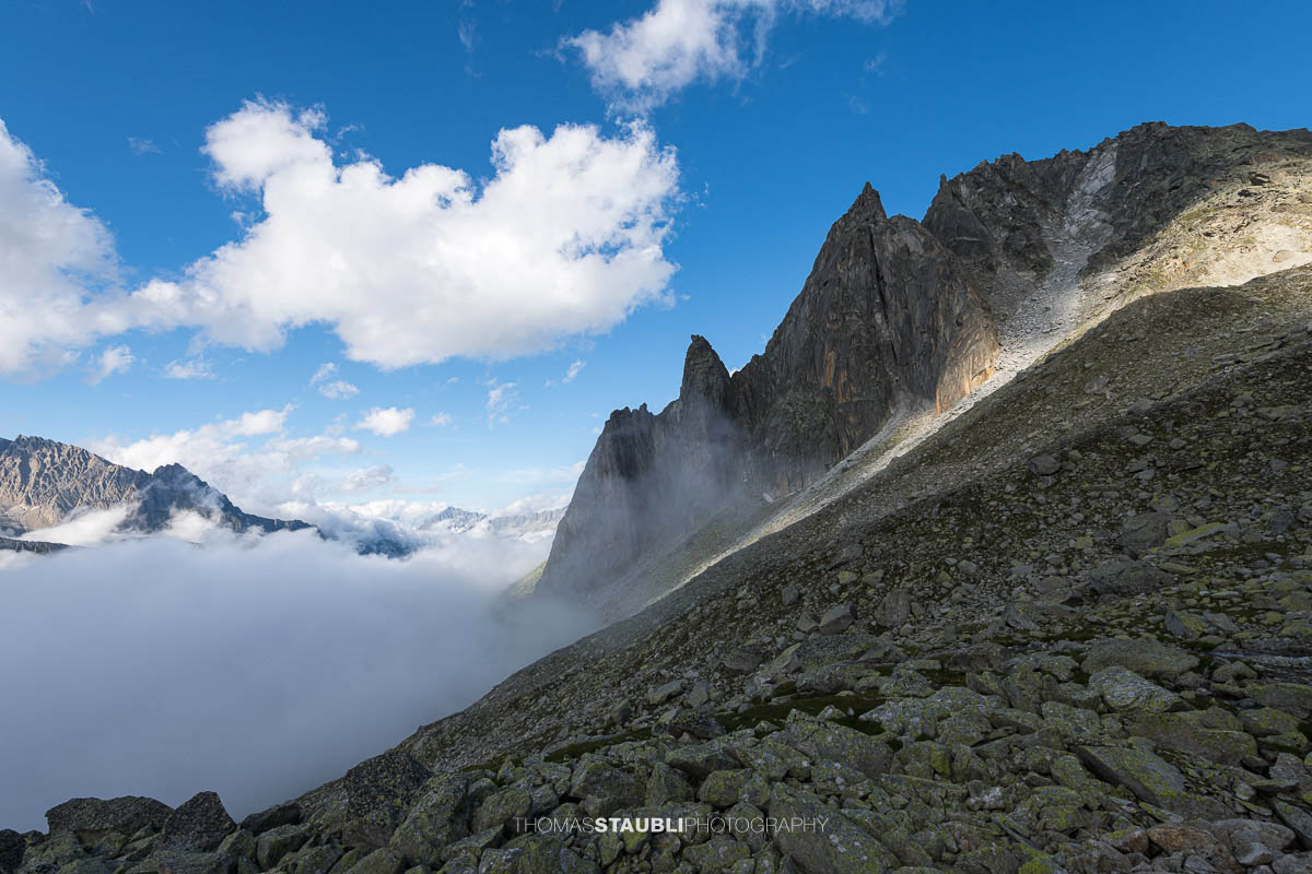 Felsige Älprigenplatten im Vordergrund, dahinter der spitze Gipfel des Feldschijen, teilweise von Nebelschwaden umgeben, unter blauem Himmel in den Urner Alpen.