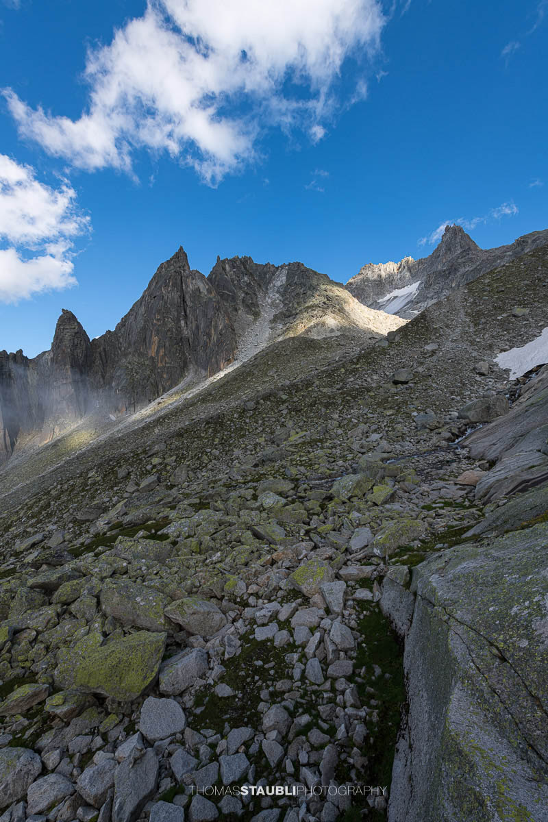 Felsige Älprigenplatten im Vordergrund, dahinter der spitze Gipfel des Feldschijen, teilweise von Nebelschwaden umgeben, unter blauem Himmel in den Urner Alpen.