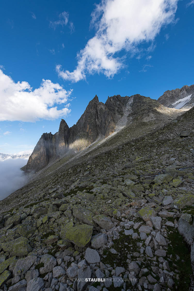 Felsige Älprigenplatten im Vordergrund, dahinter der spitze Gipfel des Feldschijen, teilweise von Nebelschwaden umgeben, unter blauem Himmel in den Urner Alpen.