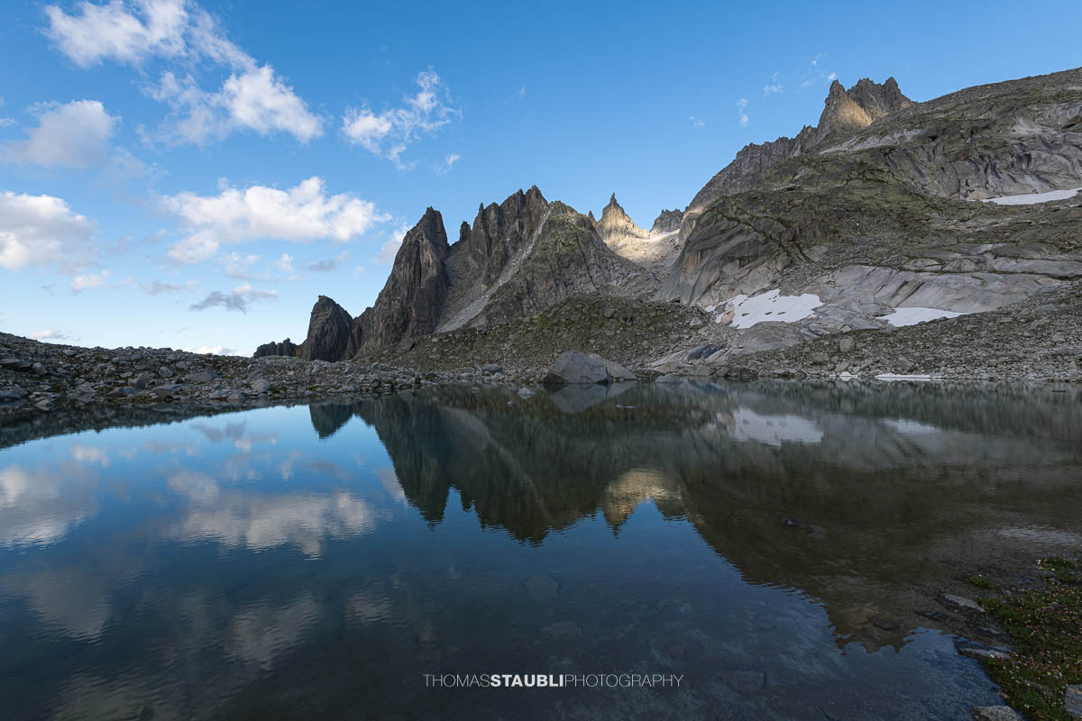 Klares Wasser des Älprigensees im Göschenertal mit Spiegelung der schroffen Felsschijen-Gipfel unter blauem Himmel.