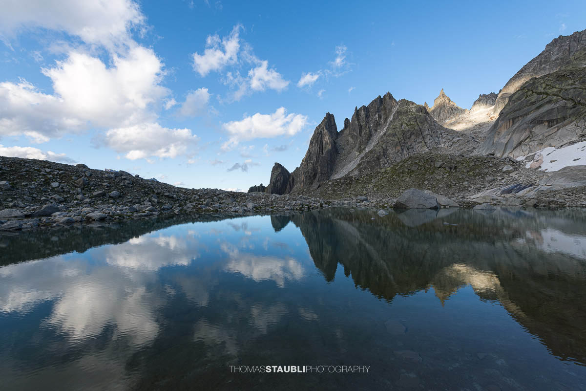 Klares Wasser des Älprigensees im Göschenertal mit Spiegelung der schroffen Felsschijen-Gipfel unter blauem Himmel.