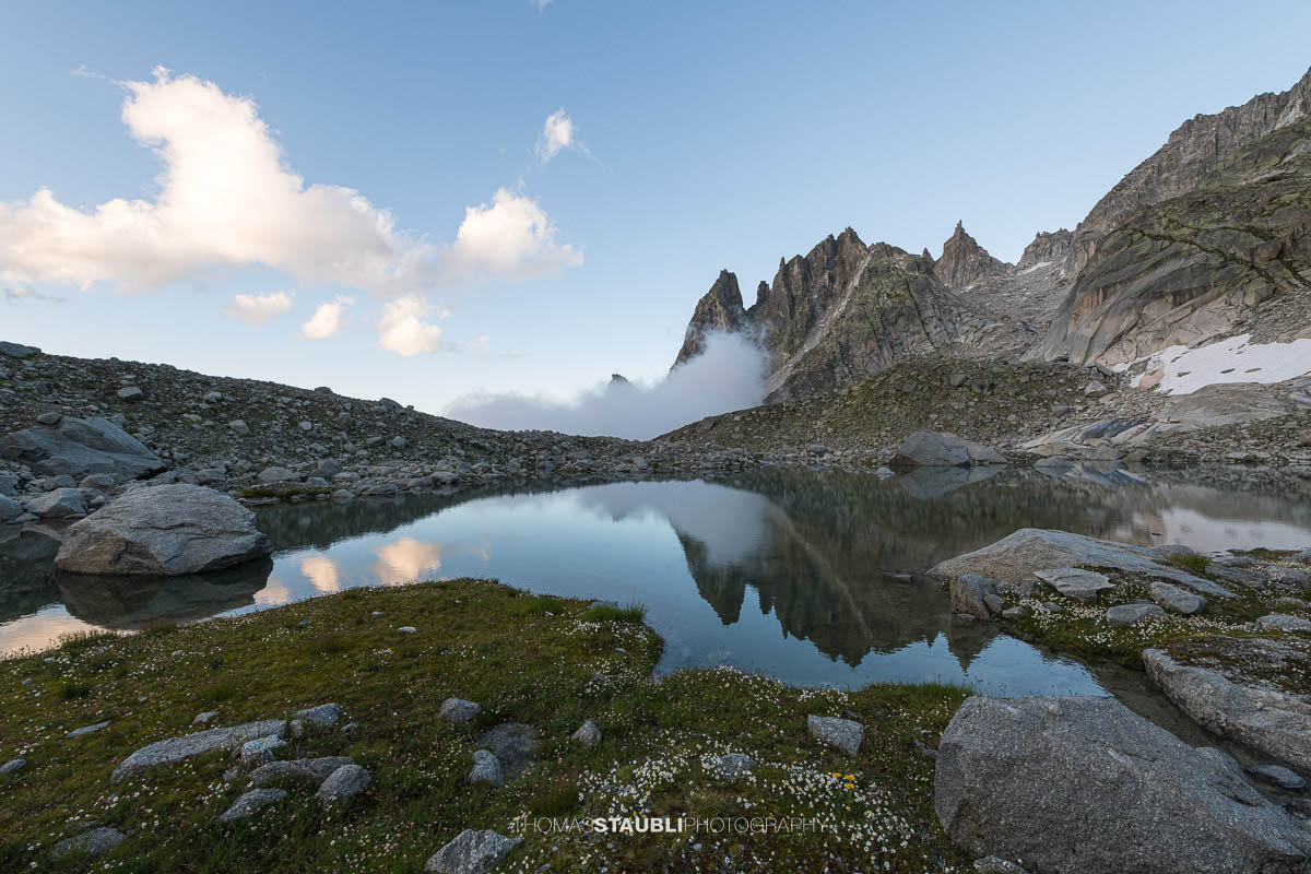 Klares Wasser des Älprigensees im Göschenertal mit Spiegelung der schroffen Felsschijen-Gipfel unter blauem Himmel.