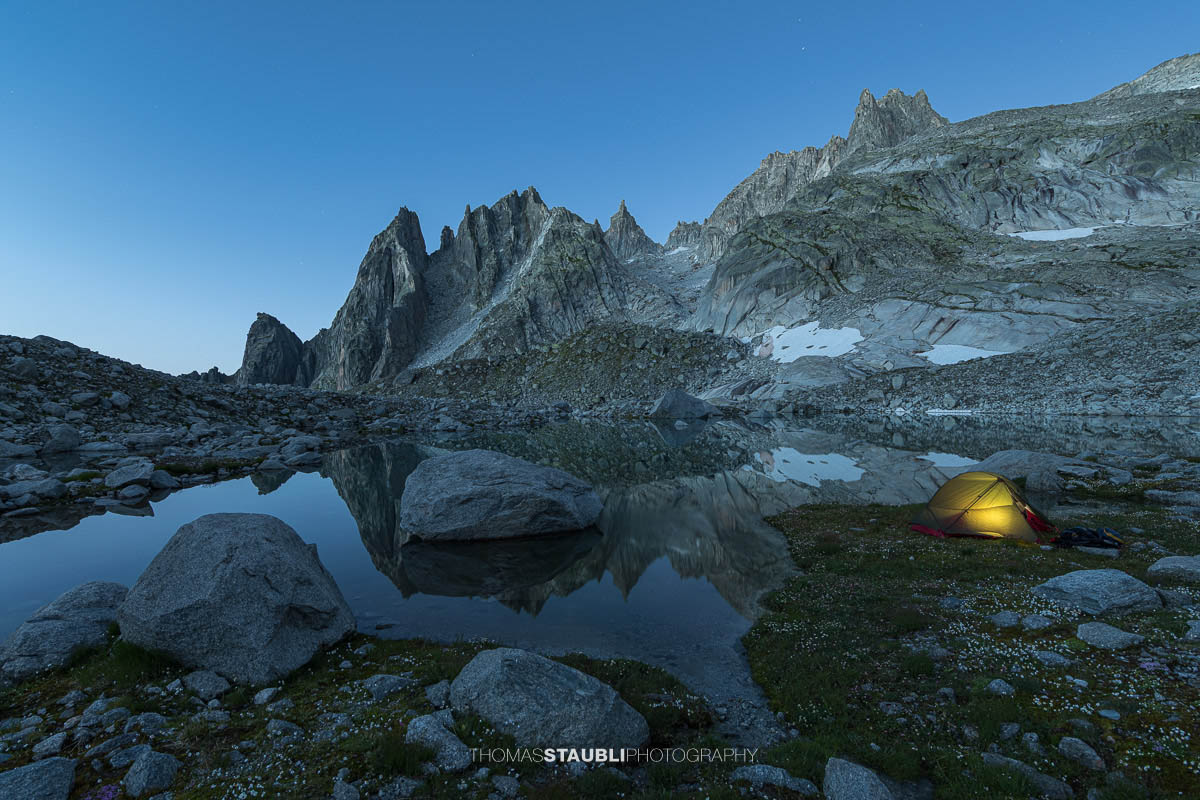 Beleuchtetes Zelt am Ufer des Älprigensees im Göschenertal bei Nacht, dahinter die schroffen Felsschijen-Gipfel im Mondlicht