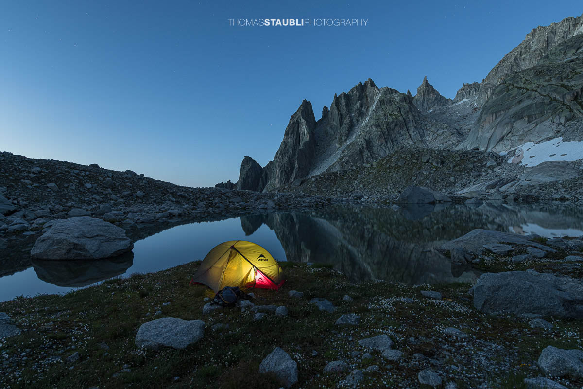Beleuchtetes Zelt am Ufer des Älprigensees im Göschenertal bei Nacht, dahinter die schroffen Felsschijen-Gipfel im Mondlicht.