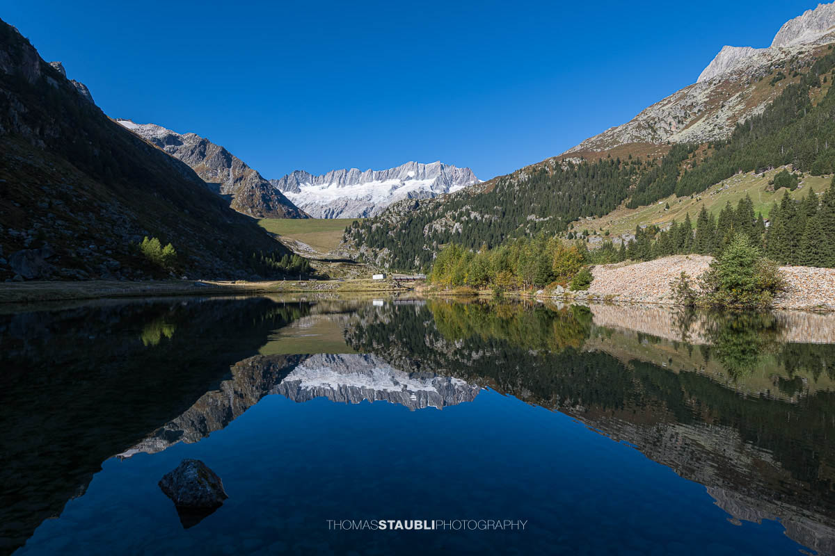 Klares Wasser im Bergsee der Göscheneralp im Kanton Uri, in dem sich die Gletscher und Gipfel des Dammastockmassivs spiegeln, umgeben von grünen Hängen und Wäldern.