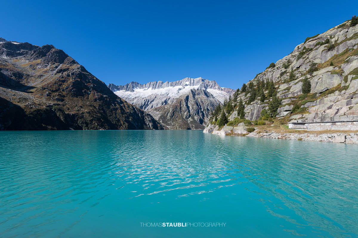 Blick über den türkisfarbenen Göscheneralpsee im Kanton Uri, umrahmt von steilen Felsflanken und den schneebedeckten Gipfeln des Dammastockmassivs im Hintergrund.