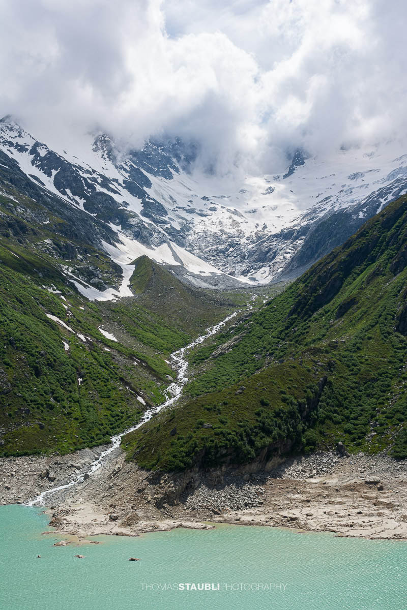 Die Dammareuss fliesst aus dem Dammagletscher hinab zur türkisfarbenen Oberfläche des Göscheneralpsees. Schneefelder, Moränen und sattgrüne Hänge prägen die wilde Hochgebirgslandschaft unterhalb des wolkenverhangenen Gletschers.