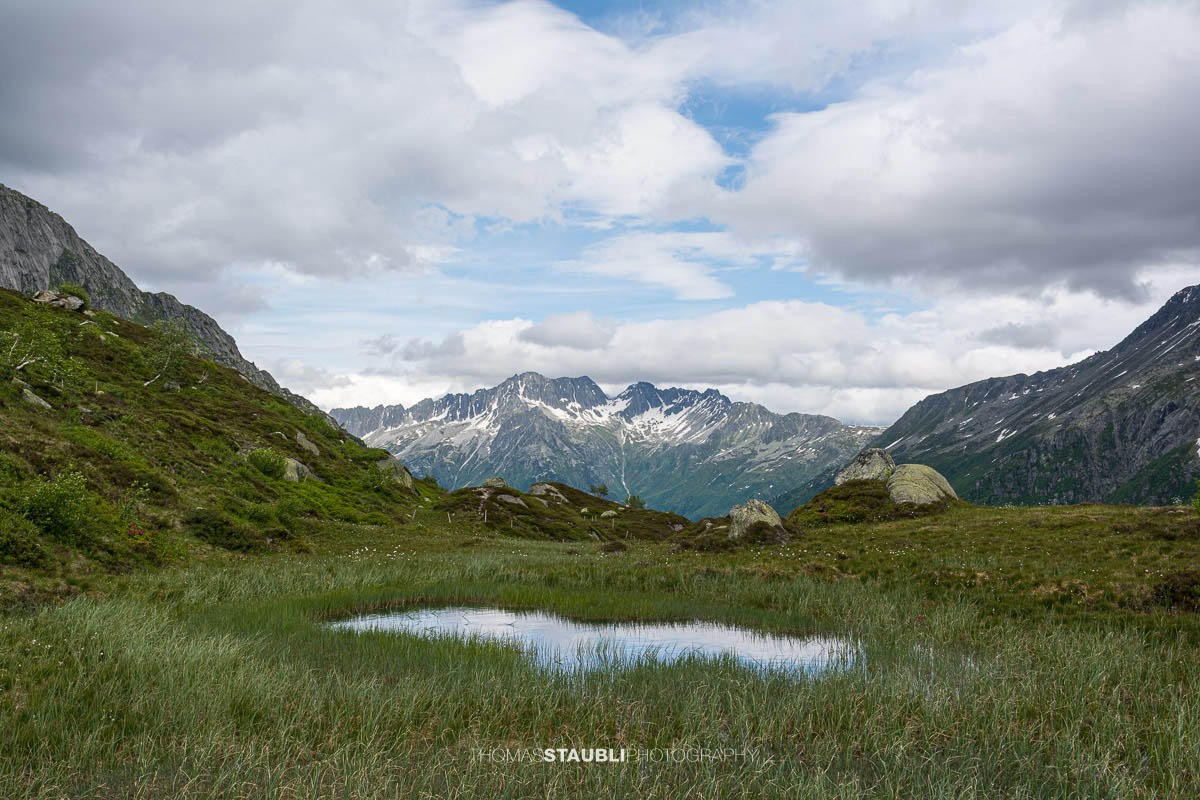 Blick talauswärts von der Göscheneralp im Kanton Uri. Ein schmaler Bergpfad führt über Felsplatten und durch alpine Vegetation, umgeben von steilen Berghängen und einer wechselhaften Wolkenstimmung.