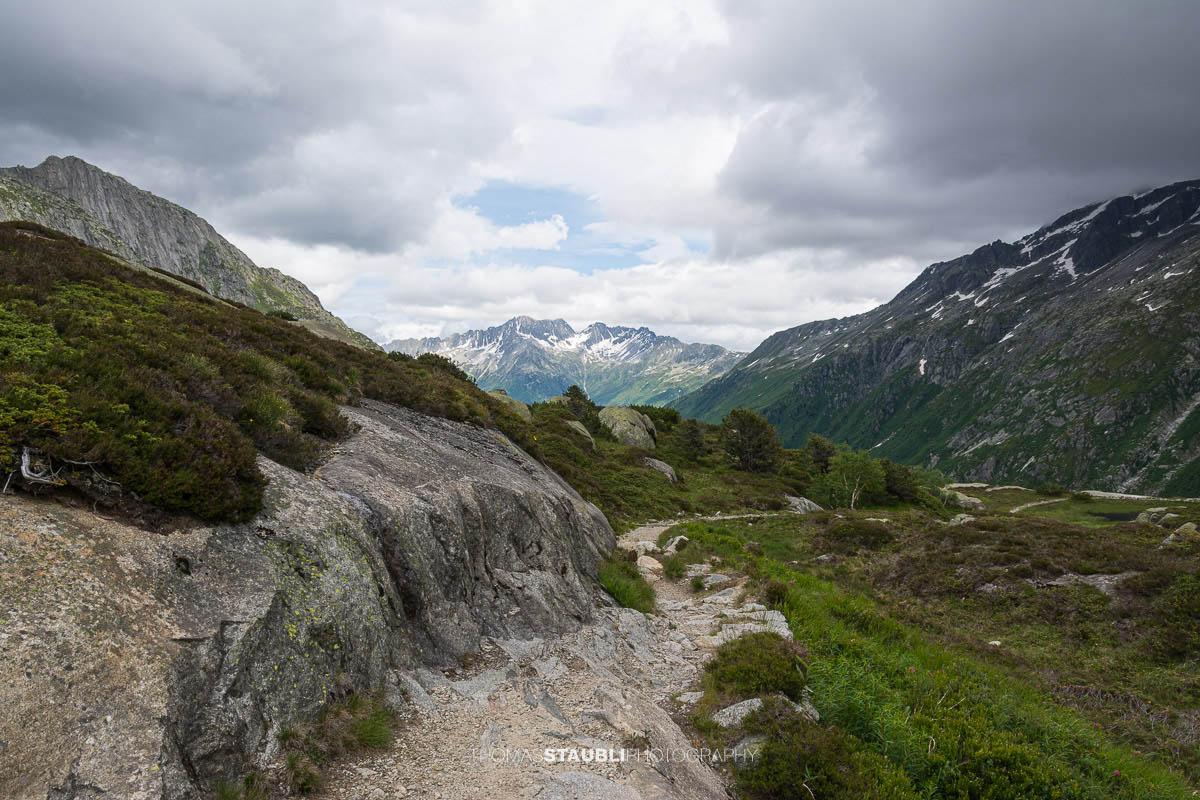 Blick talauswärts von der Göscheneralp im Kanton Uri. Ein schmaler Bergpfad führt über Felsplatten und durch alpine Vegetation, umgeben von steilen Berghängen und einer wechselhaften Wolkenstimmung.