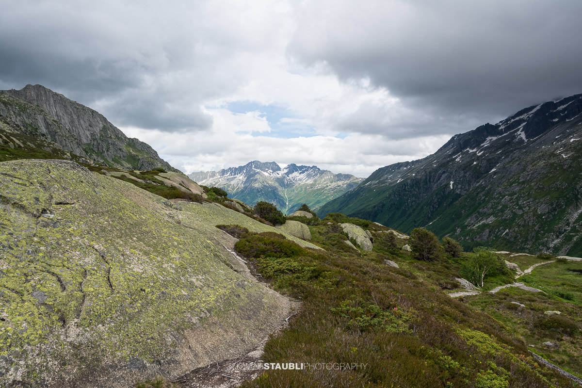 Blick talauswärts von der Göscheneralp im Kanton Uri. Ein schmaler Bergpfad führt über Felsplatten und durch alpine Vegetation, umgeben von steilen Berghängen und einer wechselhaften Wolkenstimmung.