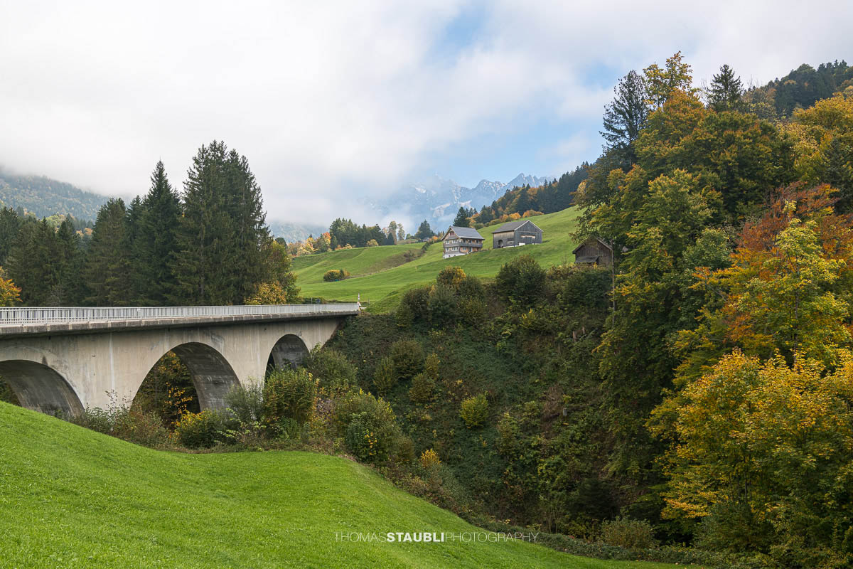 Blick vom Dorf Ennetbühl im Toggenburg an einem Herbsttag. Saftig grüne Wiesen und herbstlich gefärbte Bäume prägen die sanfte Hügellandschaft vor den traditionellen Häusern.