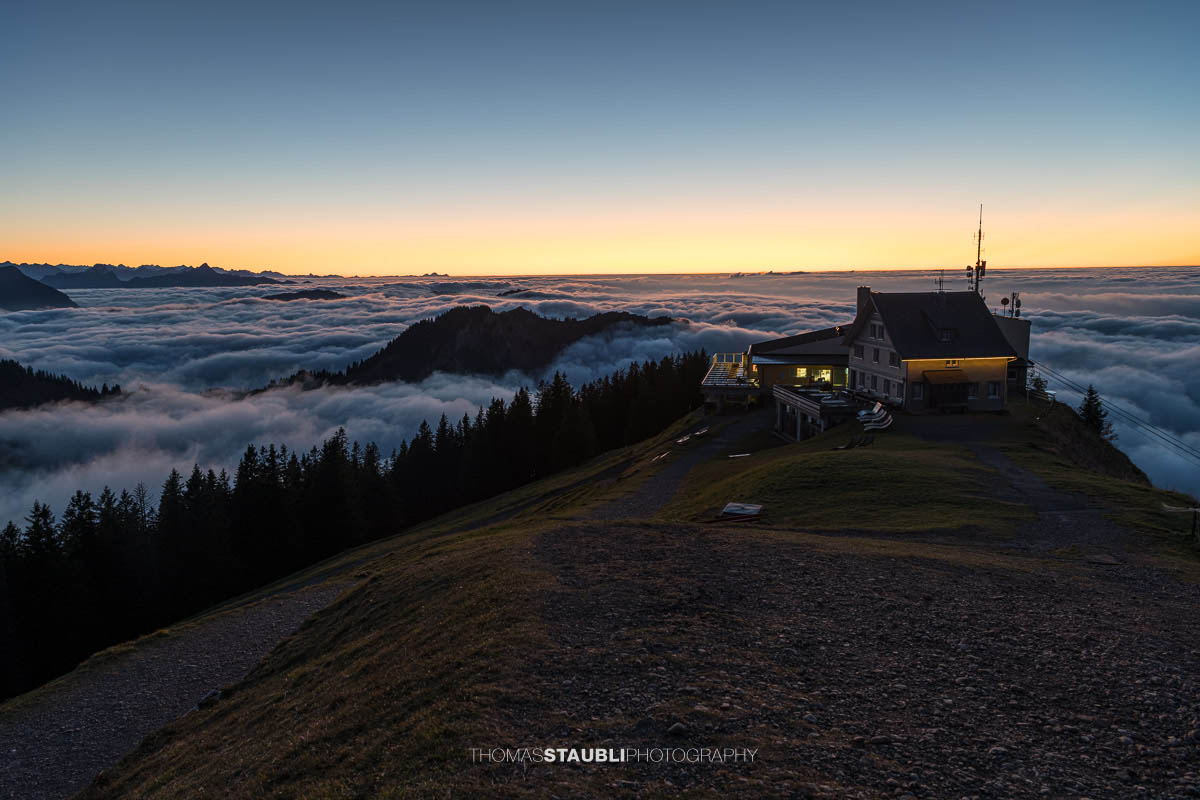 Die Kronbergbahn-Bergstation im warmen Licht der Abenddämmerung, über einem endlosen Nebelmeer mit Blick auf die fernen Alpen – eine stille und stimmungsvolle Szenerie im Appenzellerland.