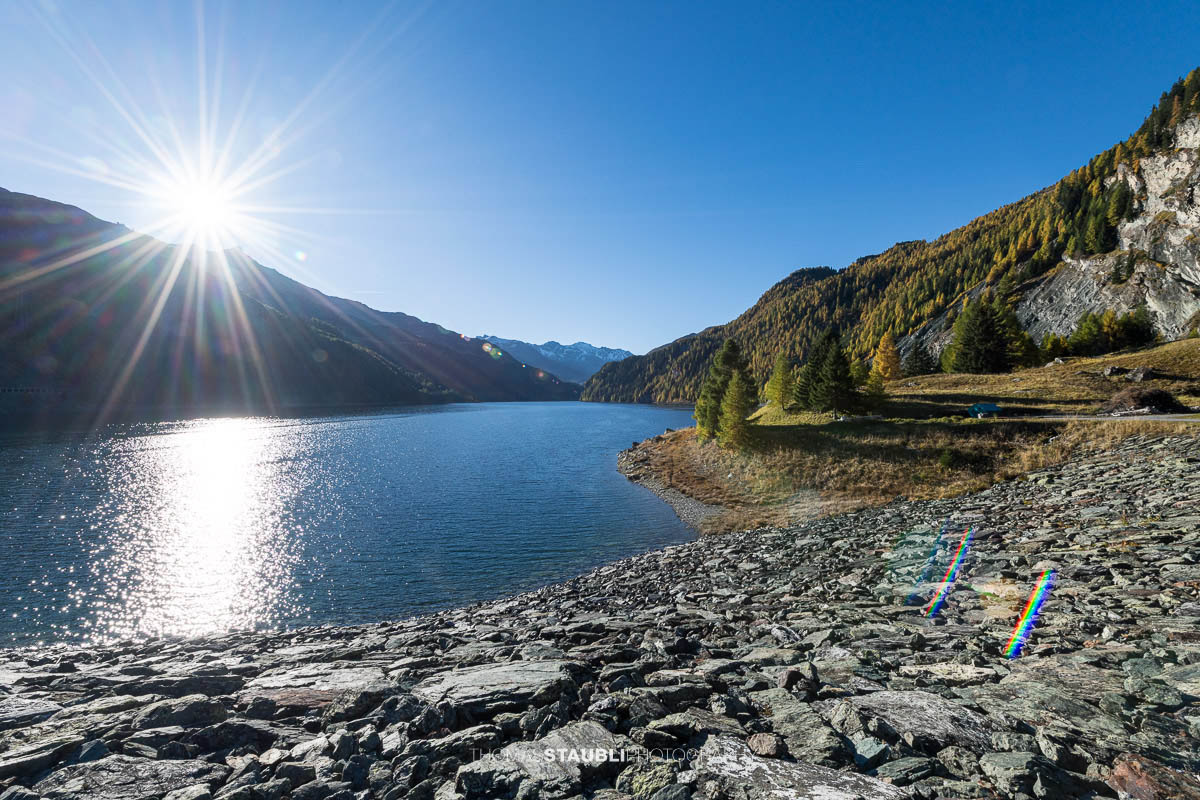 Die Morgensonne steht tief über dem Lai da Marmorera und spiegelt sich im klaren Wasser, während herbstlich gefärbte Lärchen und dunkle Felsflanken das Ufer des Bergsees im oberen Surses säumen.